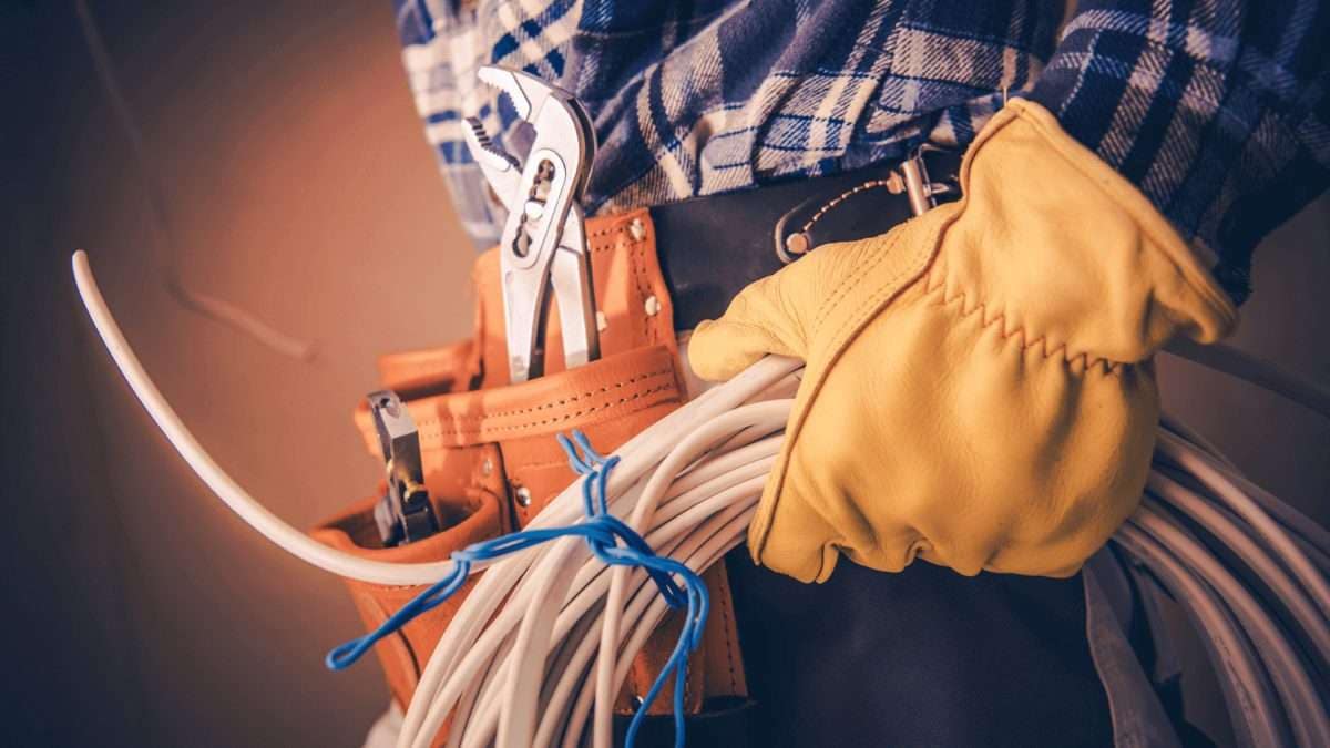 Handwerker in Arbeitskleidung mit Werkzeugen und Kabeln in der Hand.