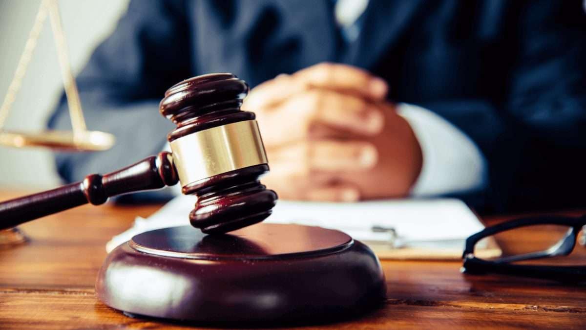 Richterhammer auf einem Tisch mit einer Person im Hintergrund, die die Hände gefaltet hat.