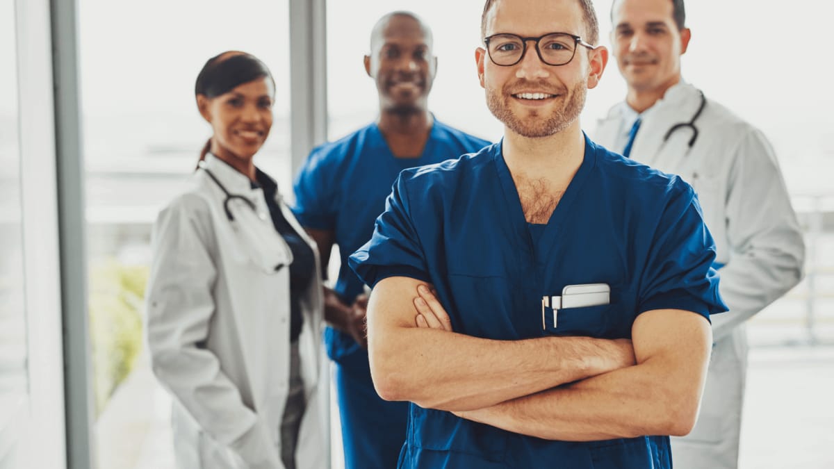 Vier medizinische Fachkräfte, darunter ein Mann in blauer Uniform und Brille, lächeln in die Kamera.