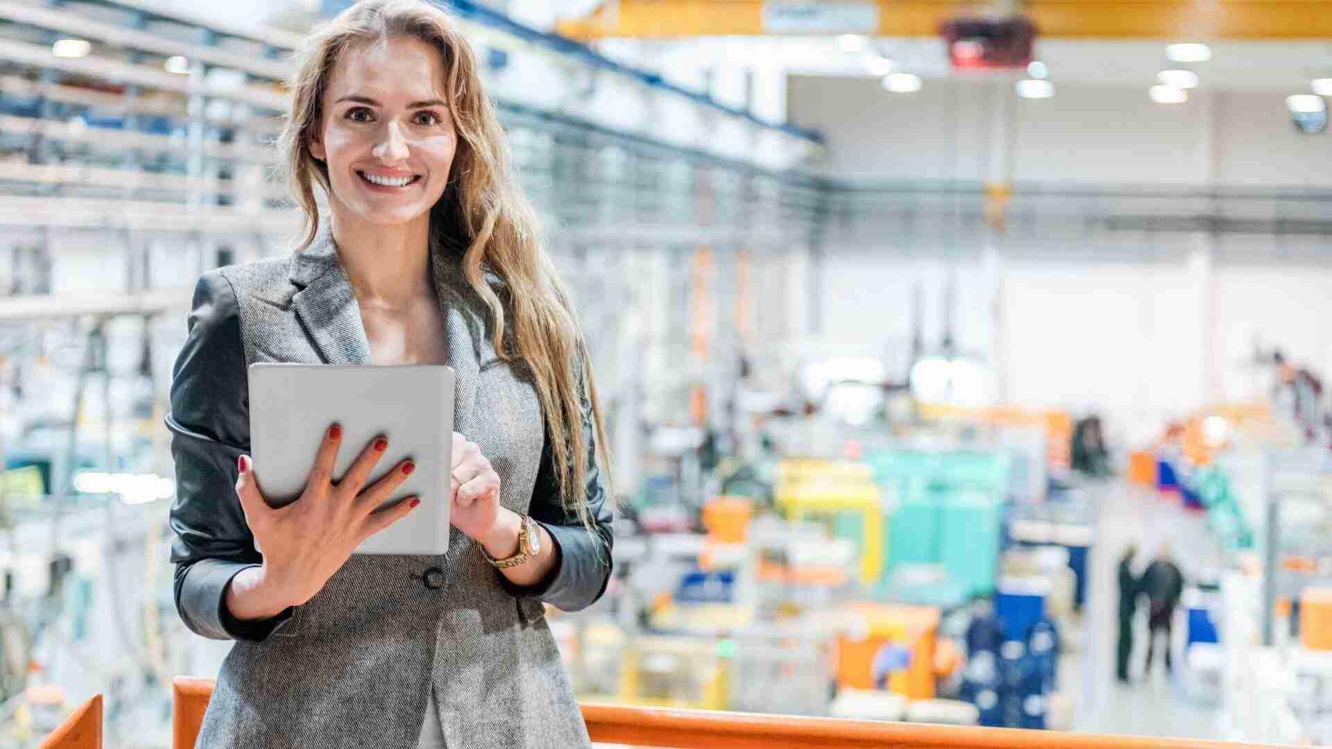 Frau mit langen Haaren hält ein Tablet in einer Fabrikhalle mit Maschinen im Hintergrund.