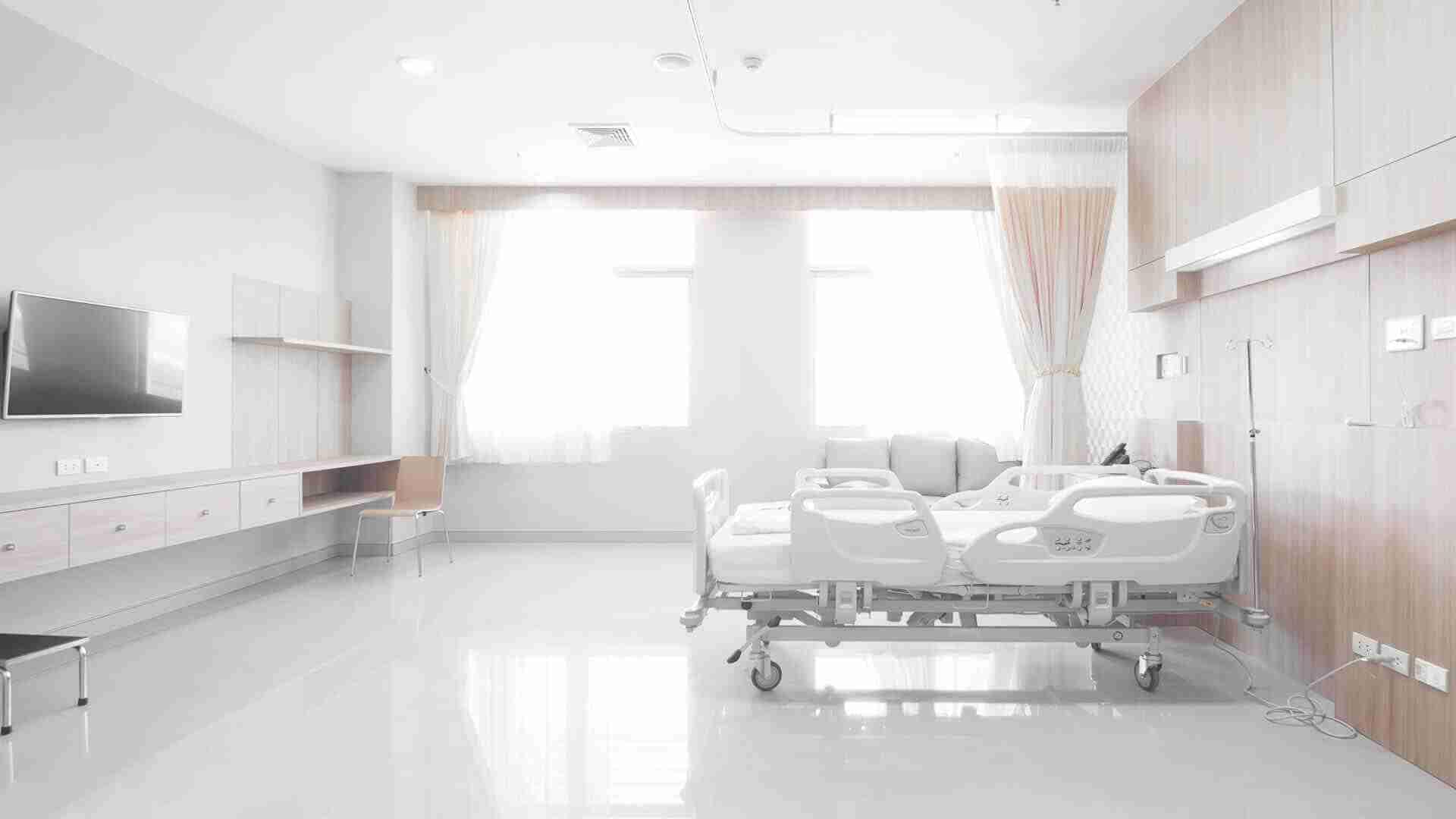 Ein Krankenhauszimmer mit einem weißen Bett, einem Fernseher und einem Fenster mit Vorhängen.