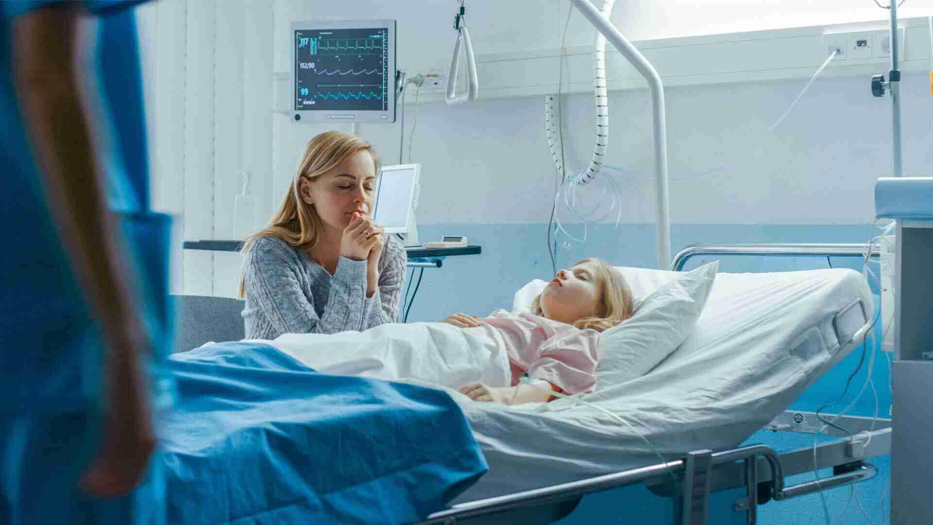 Eine Mutter sitzt am Bett ihrer Tochter, die im Krankenhaus schläft, während ein Arzt im Hintergrund steht.