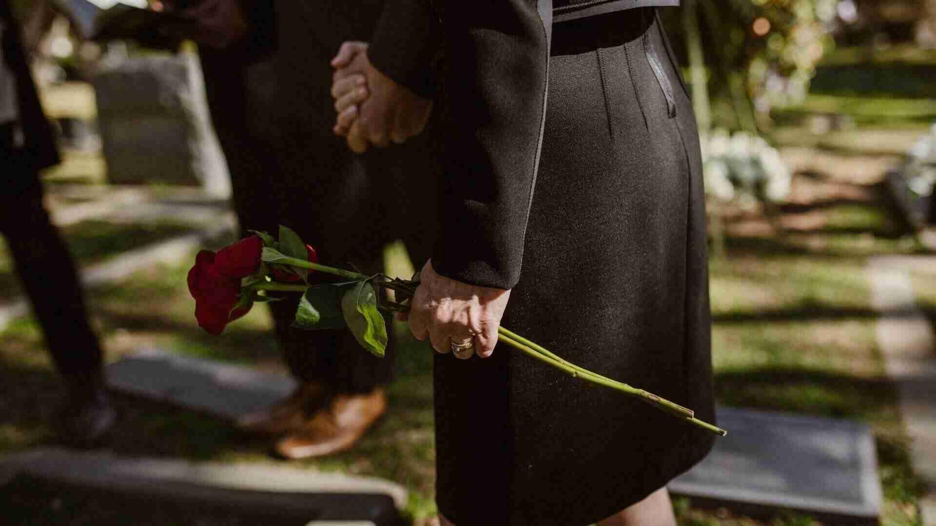 Eine Frau in schwarzer Kleidung hält eine rote Rose in der Hand auf einem Friedhof.
