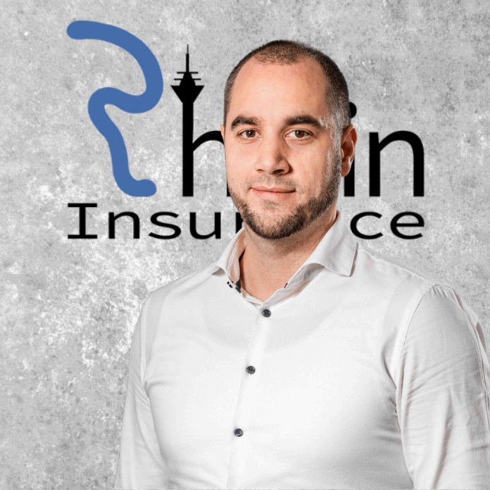 Mann mit Bart in einem weißen Hemd vor dem Logo von Rhein Insurance auf einer grauen Wand.