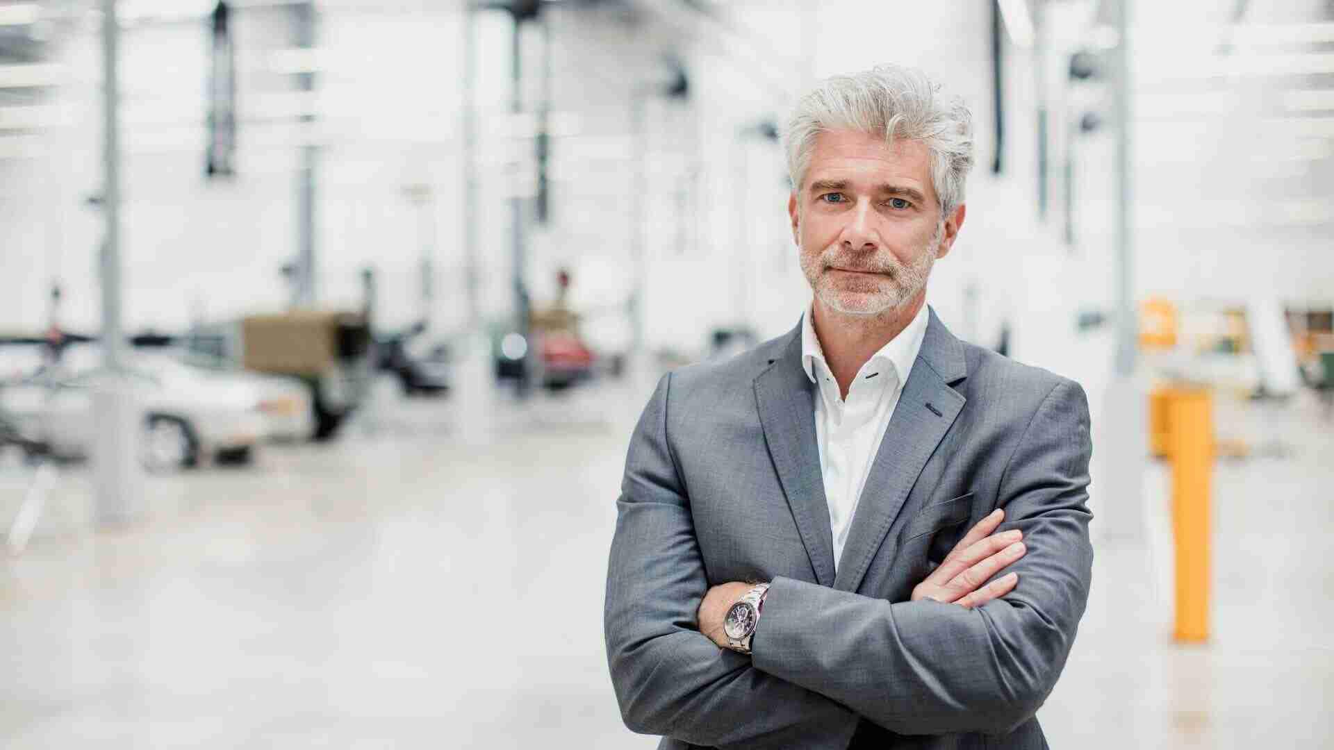 Mann mit grauen Haaren und Anzug steht mit verschränkten Armen in einer Werkstatt.
