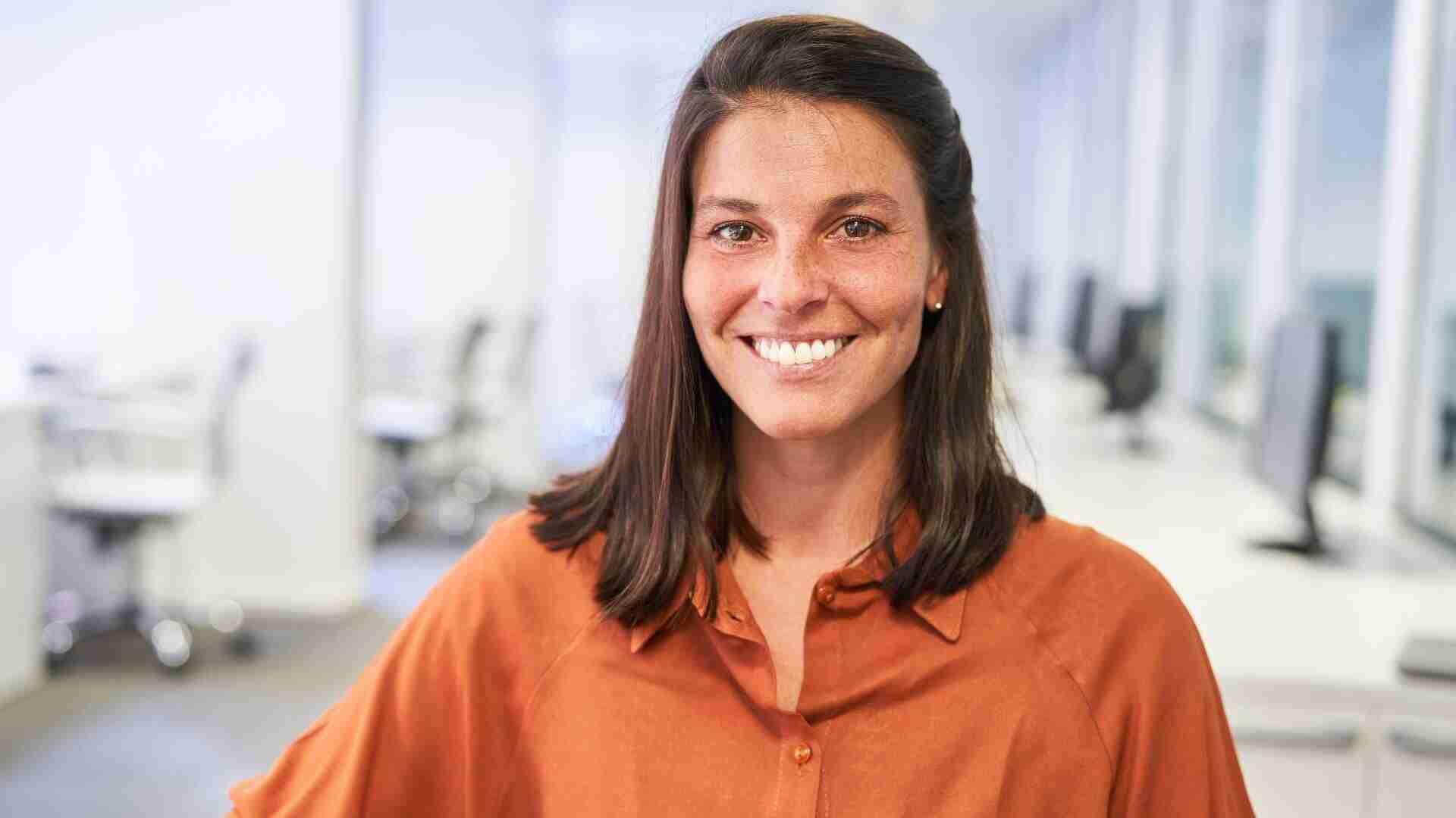Frau mit langen, braunen Haaren und einem braunen Hemd lächelt in einem Büro mit Computern im Hintergrund.