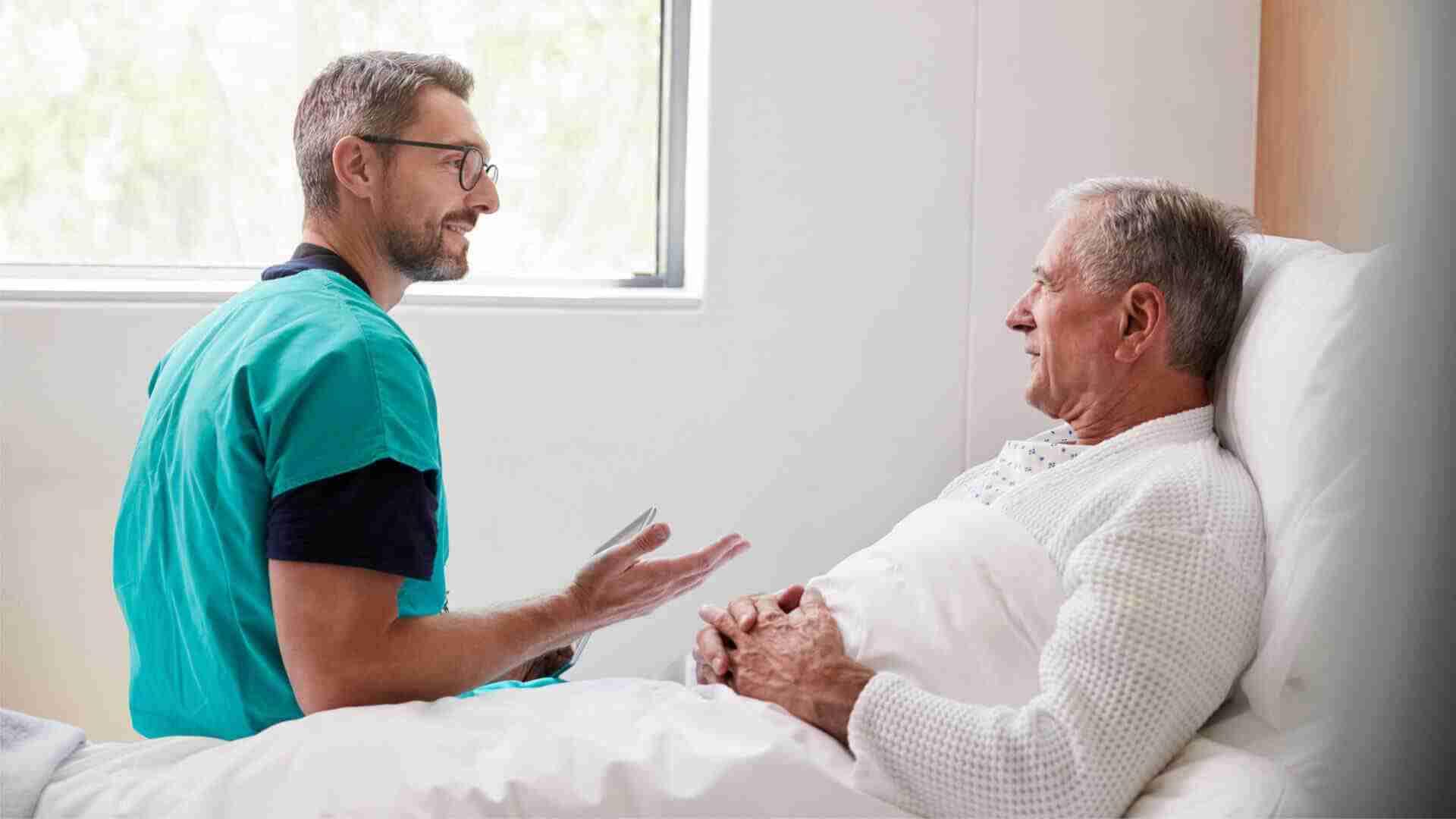 Ein Arzt spricht mit einem Patienten im Krankenhauszimmer, während der Patient im Bett liegt.