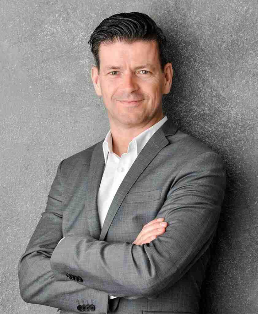 Mann mit kurzen Haaren in einem grauen Anzug und weißem Hemd, lächelt in die Kamera.