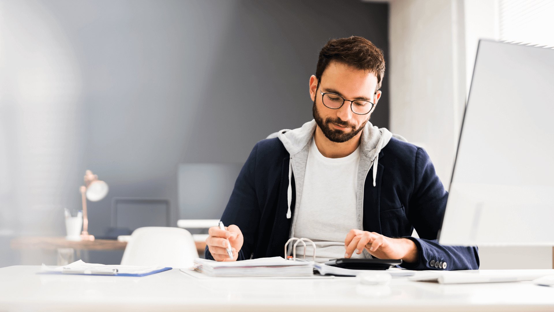 Mann mit Brille und grauem Hoodie, der an einem Schreibtisch mit Laptop und Unterlagen arbeitet.
