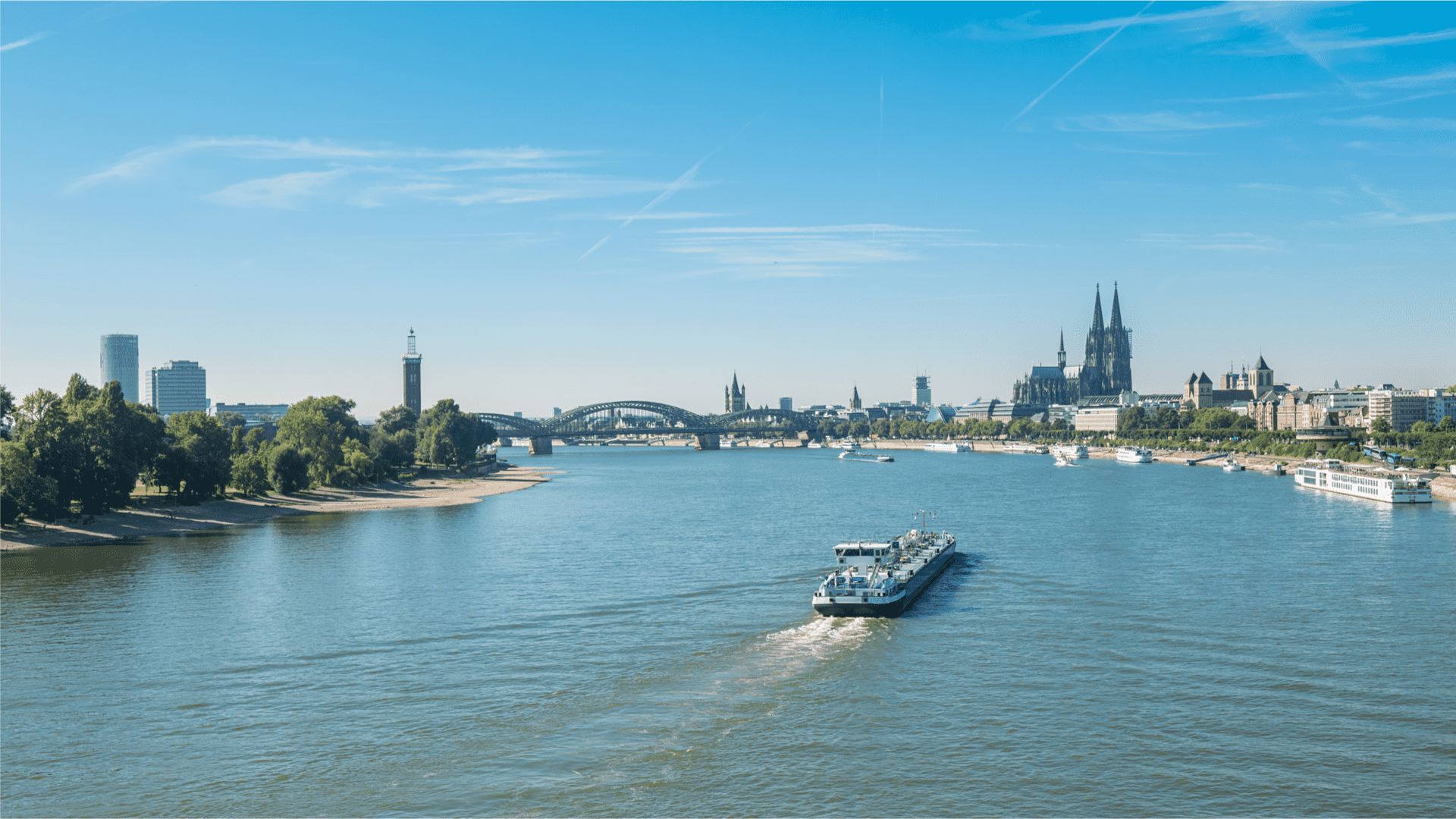 Blick auf den Rhein mit einem Schiff und der Kölner Skyline im Hintergrund an einem klaren Tag.