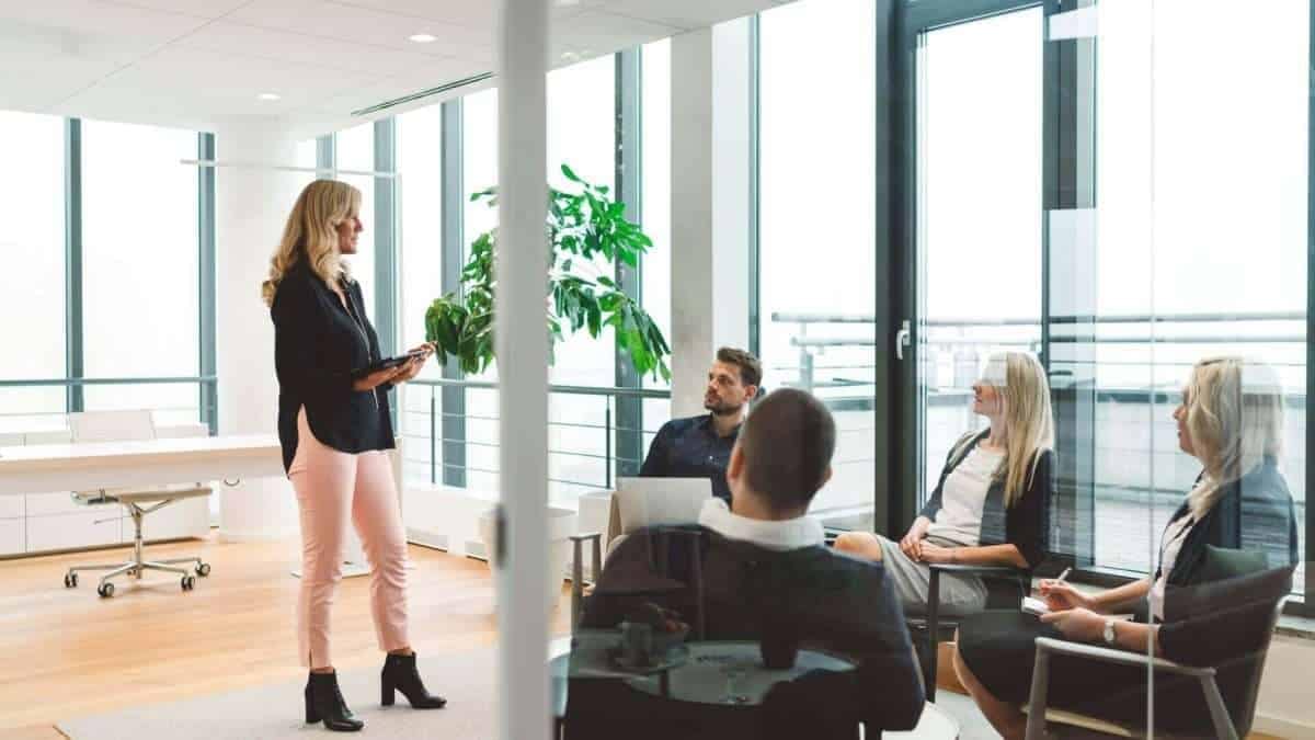 Frau steht mit einem Tablet vor einer Gruppe von Personen in einem modernen Büro.