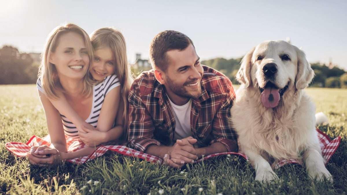 Eine Familie mit einem Hund liegt auf einer Decke im Freien und lächelt in die Kamera.