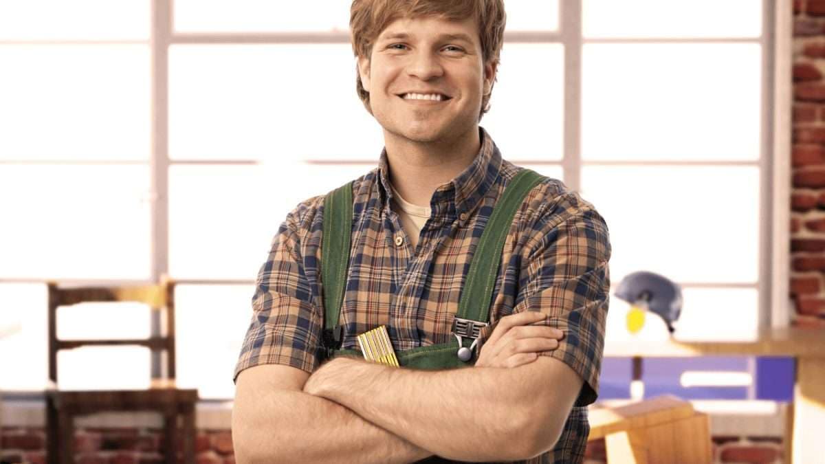 Lächelnder Handwerker in einem karierten Hemd und grüner Latzhose mit überkreuzten Armen.