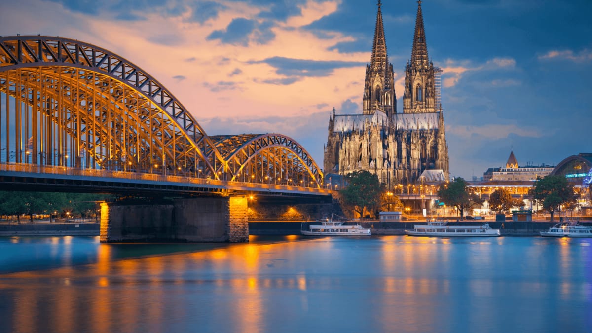 Kölner Dom und Hohenzollernbrücke mit reflektierenden Lichtern im Wasser bei Sonnenuntergang.