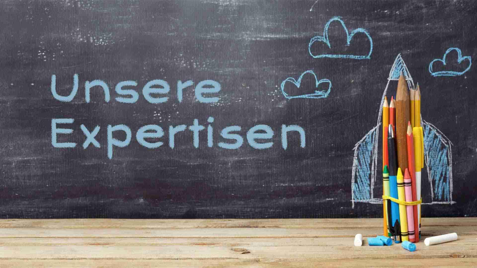 Buntstifte in einem Gummiband zusammengefasst vor einer Kreidetafel mit dem Text 'Unsere Expertisen'.