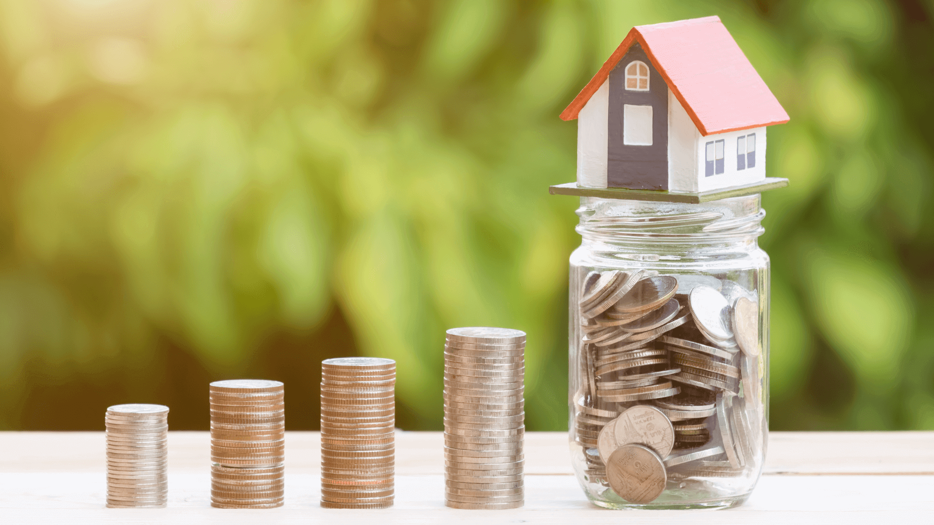 Ein Modellhaus steht auf einem Glas voller Münzen, umgeben von gestapelten Münzen.