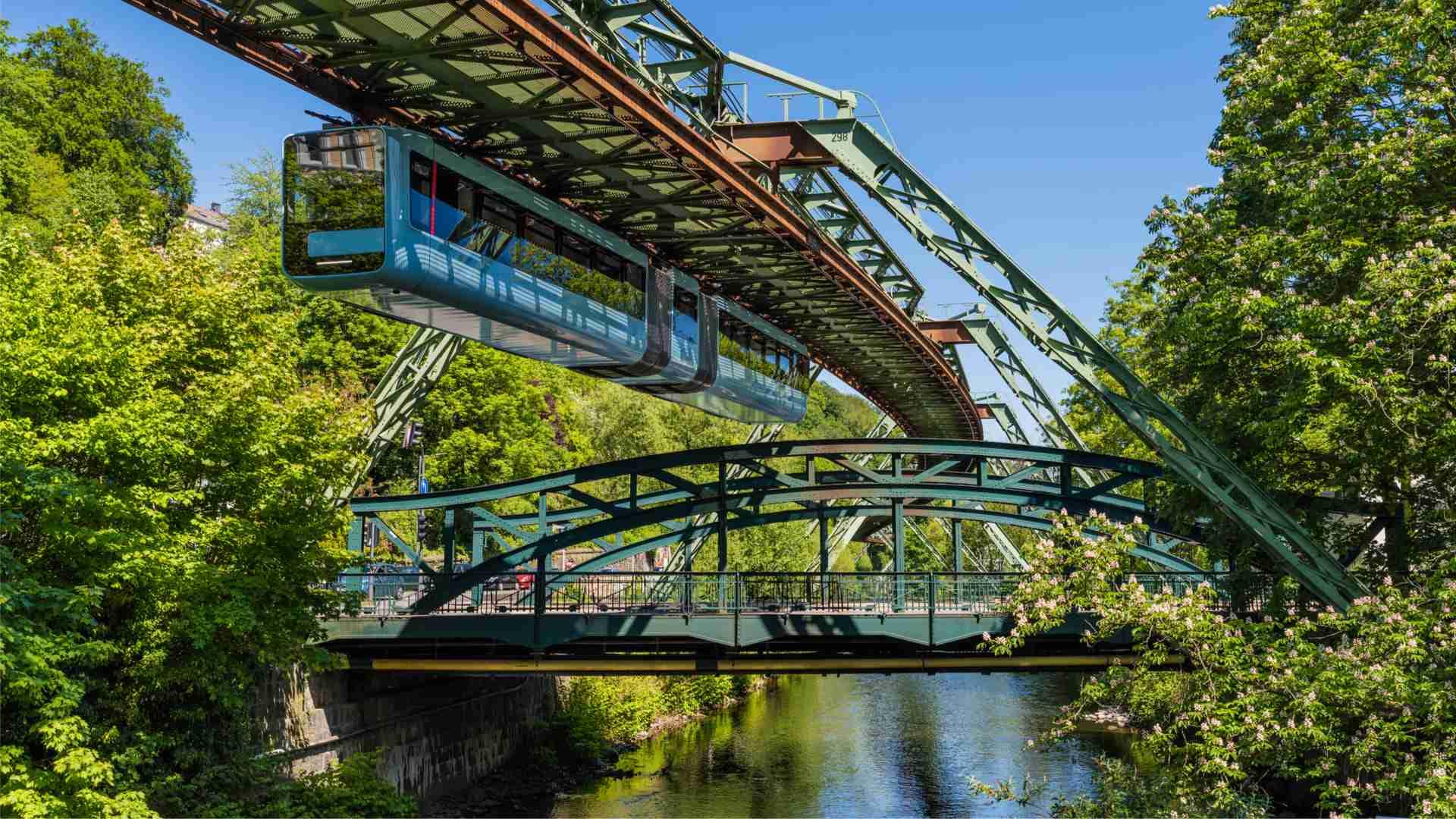 Wuppertaler Schwebebahn, eine hängende Straßenbahn, die über eine grüne Brücke und einen Fluss fährt.