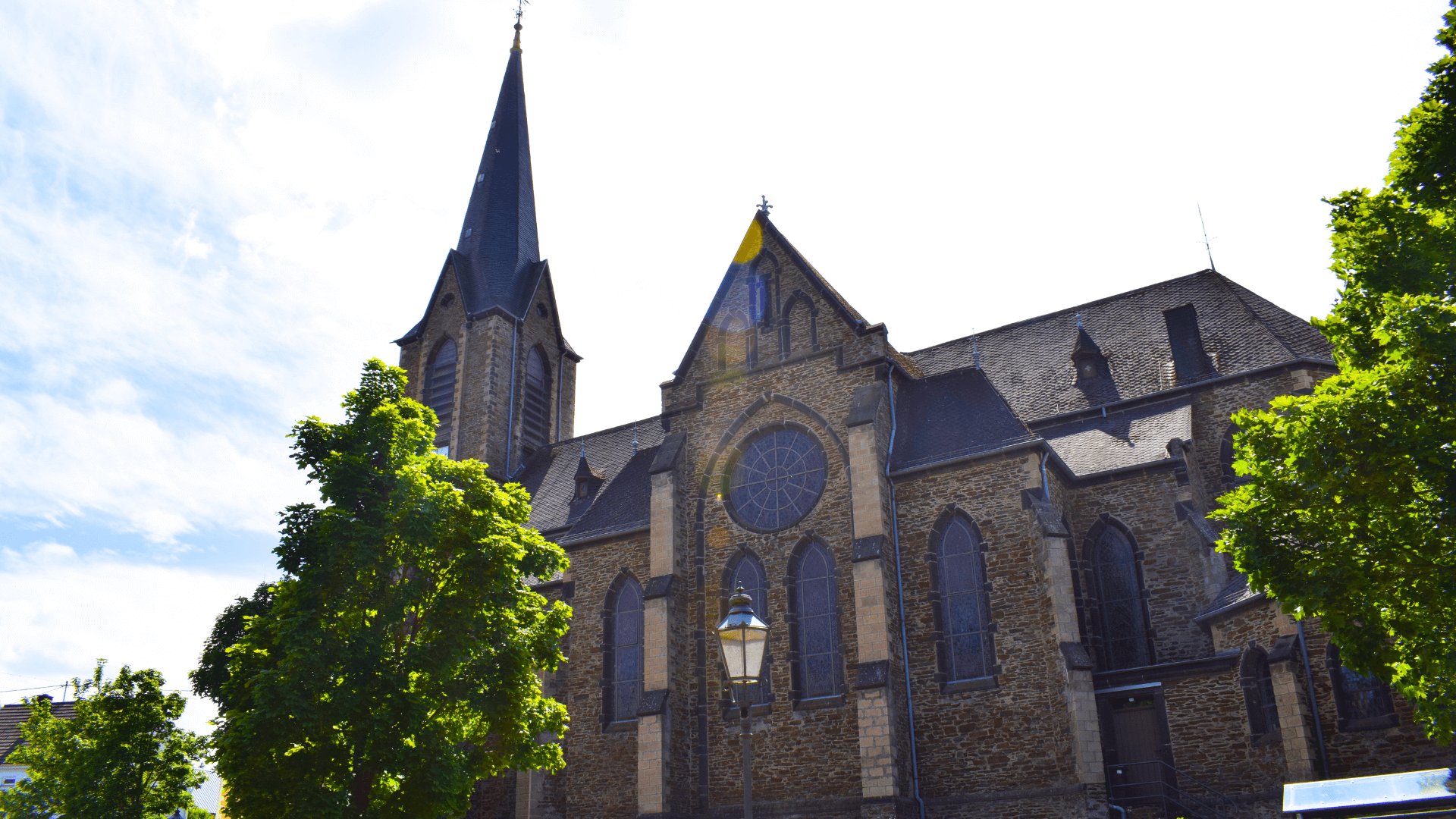 Kirche mit spitzem Turm und großen Fenstern, umgeben von Bäumen in einer städtischen Umgebung.