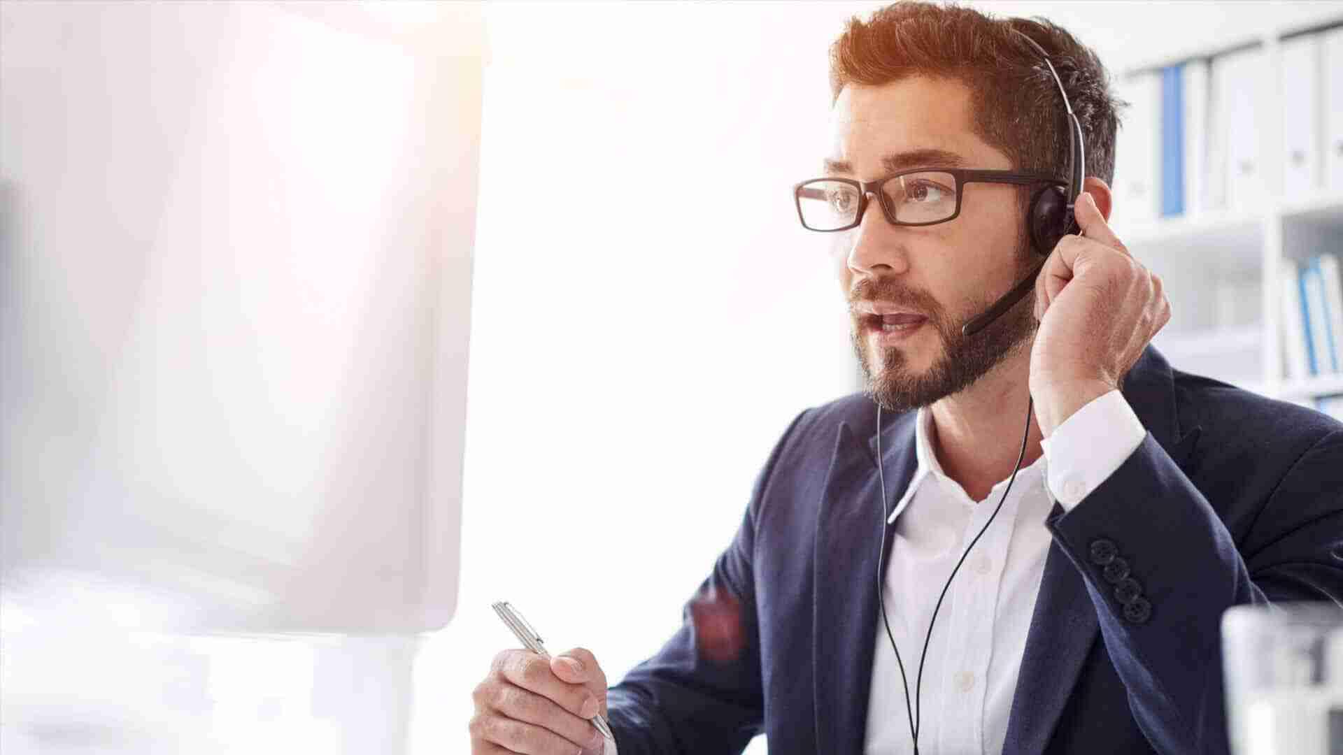 Mann mit Brille und Headset, der am Computer arbeitet und Notizen macht.