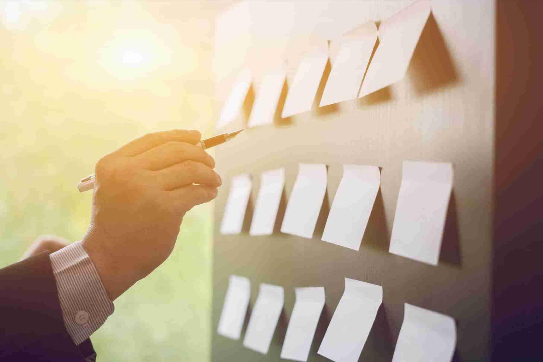 Eine Hand, die einen Stift auf ein leeres Notizblatt an einer Wand hält, mit sanftem Licht im Hintergrund.