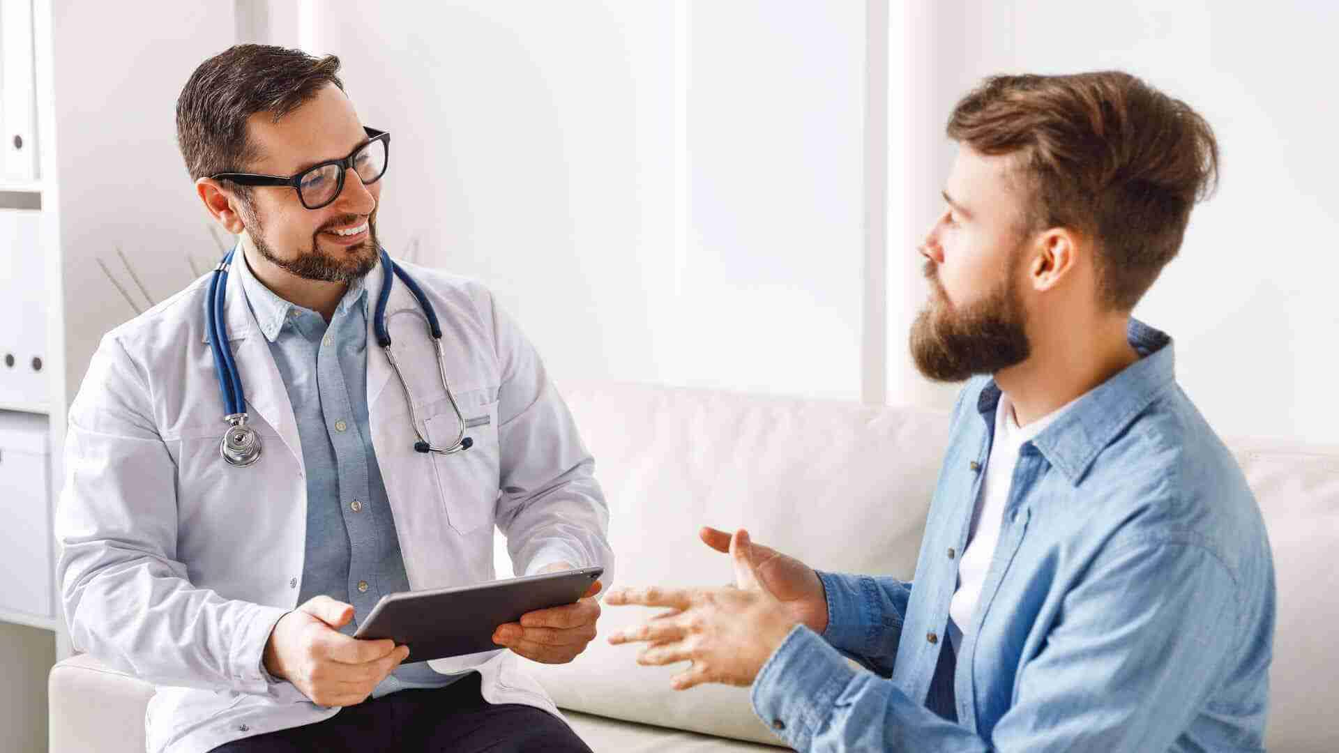 Ein Arzt mit Stethoskop spricht mit einem Patienten, der auf einem Sofa sitzt und gestikuliert.